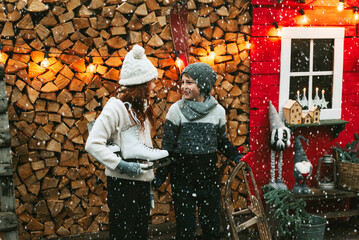 siblings teenage girl sister and cute boy brother in knitted sweater and hat stand at porch of country house with sleds and skates, concept of winter sports and Christmas holidays for children outdoor