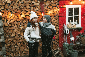 Obraz premium siblings teenage girl sister and cute boy brother in knitted sweater and hat stand at porch of country house with sleds and skates, concept of winter sports and Christmas holidays for children outdoor