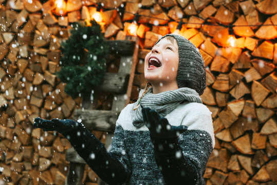 cute boy brother in knitted sweater and hat having fun with first snow and catches snowflakes at porch of country house, concept of winter sports and Christmas holidays for children outdoor