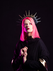 A gothic diva with pink hair in a royal image, a crown with roses and rays on her head, hands folded in a prayer gesture.