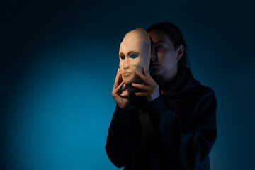 Hiding behind a mask, a young woman in a dark hoodie hides her face with a mask, the concept of self-doubt. Portrait in the studio on a dark gray background.