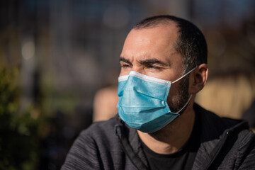 Young male or man wearing a surgical mask in public space sitting behind a table. Casual sunny day with covid protective mask on.