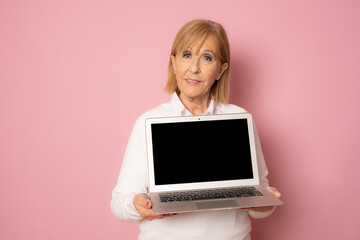 Portrait of a cheerful mature woman showing blank screen laptop computer isolated over pink...