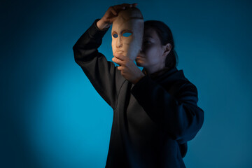 Hiding behind a mask, a young woman in a dark hoodie hides her face with a mask, the concept of self-doubt. Portrait in the studio on a dark gray background.