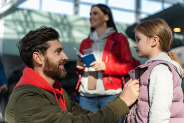 Happy family with little kid traveling by airplane. Father straightens clothes to his daughter while sitting at the airport. Mother standing at the background. Traveling concept