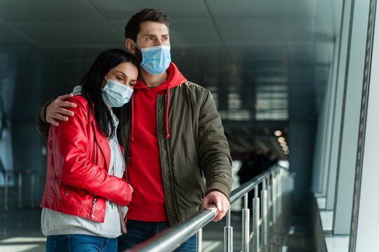 Sad Couple In Love Wearing Protective Mask Embracing At The Airport Hall While Standing In Front Of The Window And Waiting For Something