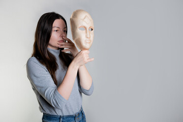 Complexes due to appearance or self-identification problems, concept. A young woman holds a mask in her hands hiding her face. Photos in light gray tones, gray background