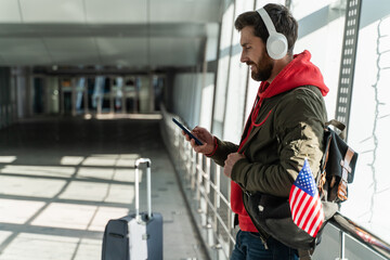 Close up view of the traveler man wearing headphones looking at the smartphone screen while listening music and waiting for his flight. Citizens and american symbols concept concept