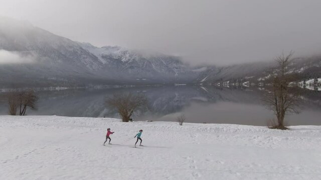 AERIAL: Flying along two female athletes training nordic skiing in Slovenian mountains. Two young women train nordic skiing around the spectacular lake Bohinj. Drone shot of people skiing around lake.