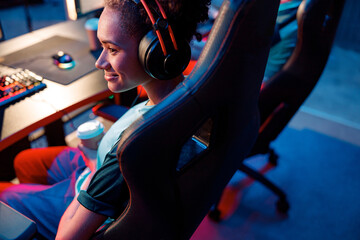 Cute African woman wearing headphones sitting on gaming chair and holding coffee cup during online esports tournament in computer club