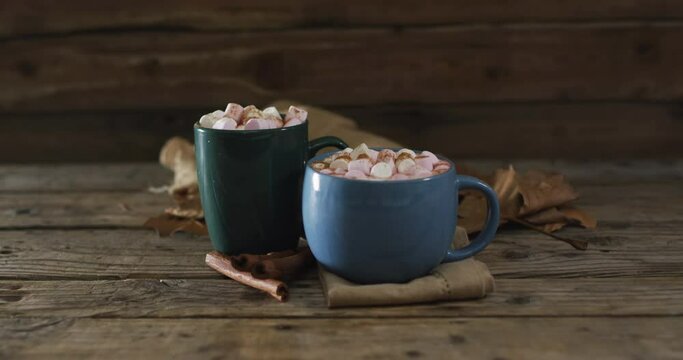 Two cups of hot chocolate with marshmallows, autumn leaves and cinnamon sticks on wooden surface