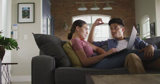 Happy biracial couple sitting on sofa in living room, using laptop