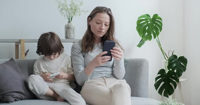 Focused modern mother and son kid chatting surfing internet use smartphone sitting on couch together. Confident family suffering social networks mobile phone addiction spending time procrastination