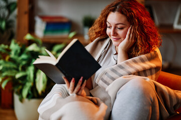 A beautiful ginger girl sitting in the living room wrapped in a blanket and reading an interesting...