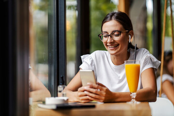 A happy young woman sits in a bar with wireless headphones on and hanging on the social network. A woman takes a break from fast living. A woman in a cafe with a phone