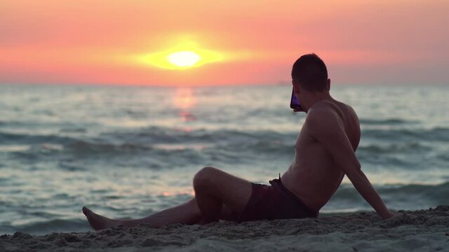 Young man sitting on the tropical sandy beach and taking photos of the beautiful beach sunset with smart phone