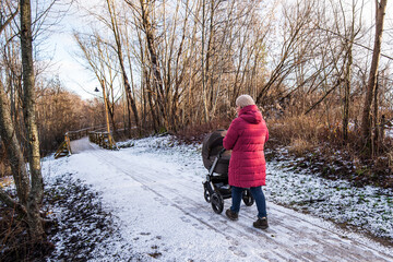 Fototapeta na wymiar Woman with a pram walking along the walking path. First snow and footprints, wooden bridge, pole with lantern on sunny autumn day. Kuldiga, Latvia