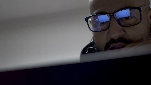 A close up shot of an Indian man talking into his microphone headset during a conference call, the light from the computer screen reflecting off the tint of his glasses as he reads the details