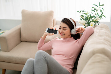 Happy young Asian woman in earphones singing and listening to music while sitting on sofa and holding mobile phone at home.