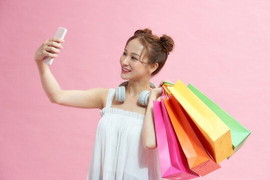 Happy young woman with shopping bags taking selfie on pink background
