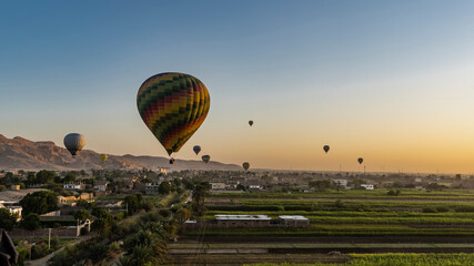 Colorful balloons fly at dawn over the Nile Valley. Below are cultivated fields, village houses. A mountain range against the background of the sky, highlighted in orange. Luxor