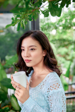 Hanging Potted Plants In Summer With Woman Standing On Porch Of House In Morning Wooden Cabin Cottage Drinking Tea Or Coffee