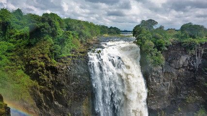 Obraz premium The Zambezi River flows along the plateau and collapses with a powerful Victoria Falls stream. Rainbow over the gorge. Lush green vegetation around. Cloudy sky. Zimbabwe