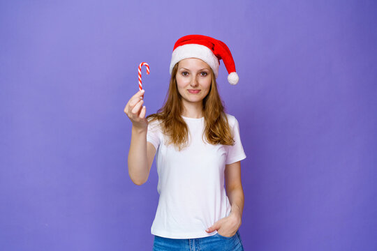 Portrait of woman in santa hat and white t-shirt, a girl holding a cane-shaped lollipop on a lilac background. Caucasian young woman with sweet tooth