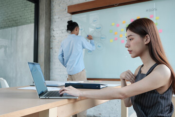 Two colleagues and coworkers of Asian ethnicity brainstorm and meeting finance project discuss with business plan in conference room with colorful sticky notes paper on writing board in the office.