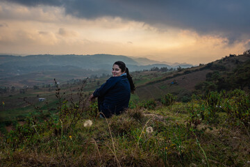 young girl sitting on mountain top with misty hill range background and dramatic sky at morning