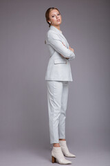 Fashion photo of a beautiful elegant young woman in a pretty soft blue suit, jacket, pants, trousers, white boots posing on gray background. Studio shot, portrait. Monochrome colors