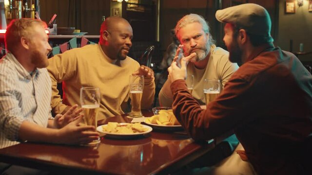Group of four stylish bearded men sitting together in modern pub drinking beer, eating snacks and chatting