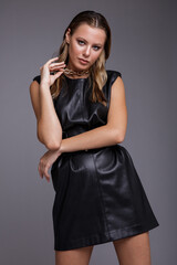 Fashion photo of a beautiful elegant young woman in a pretty leather dress, bronze-peach metallic boots, thick metal chain around her neck posing over gray background. Studio shot, portrait. 