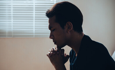 A faithful Asian young male hands in prayer gesture sitting alone on sofa at home office pray and...