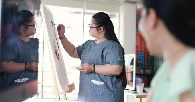 Side view of Asian female teacher talking to girl with down syndrome creating painting on canvas during art lesson