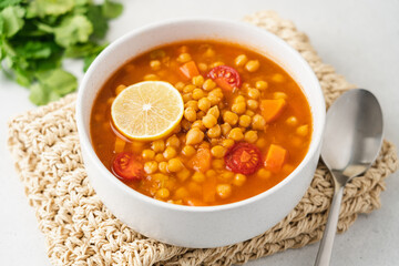 Chickpea lentil soup with vegetables, cilantro and lemon in light bowl on white table. Vegetarian soup side view