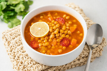 Chickpea lentil soup with vegetables, cilantro and lemon in light bowl on white table. Vegetarian...