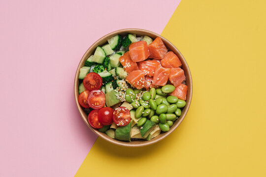 Salmon, avocado, cucumber, tomato, edamame beans and rice poke bowl on pink yellow background