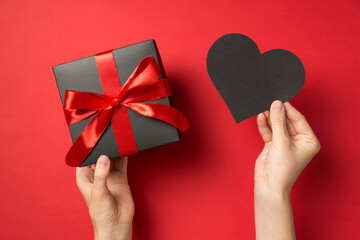 First person top view photo of hands holding black paper heart and black giftbox with red ribbon...