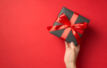 First person top view photo of girl's hand holding black giftbox with red ribbon bow on isolated red background with empty space