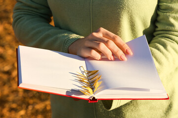 Woman holding opened book with autumn leaf on sunny day, closeup