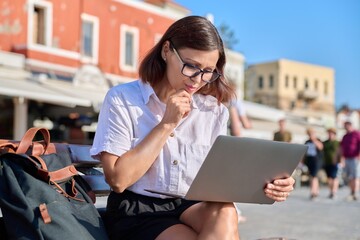 Middle-aged woman sitting on a bench in city, using a laptop
