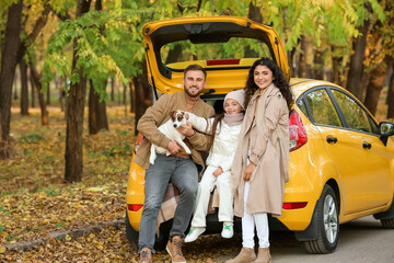 Happy parents with little daughter and cute dog sitting in car trunk on autumn day