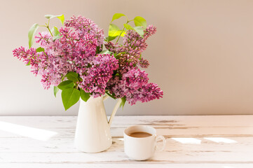 fresh branches of lilacs in a jug on a white wooden table with a cup of shaded coffee.