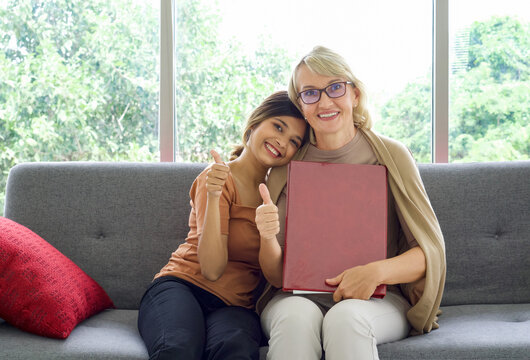 Asian daughter and her adopted caucasian mother raise finger thumbs up while holding red leather document folder. Happy mix skin family living together.