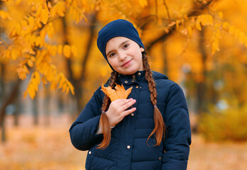 Portrait of a child girl in autumn city park. Posing with maple leaf. Beautiful nature, trees with yellow leaves.