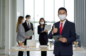 Asian businessman in suit lifted a finger thumb up while holding tablet computer. The team is preparing for the meeting in the background. Everyone wears a mask. Concept of leadership.