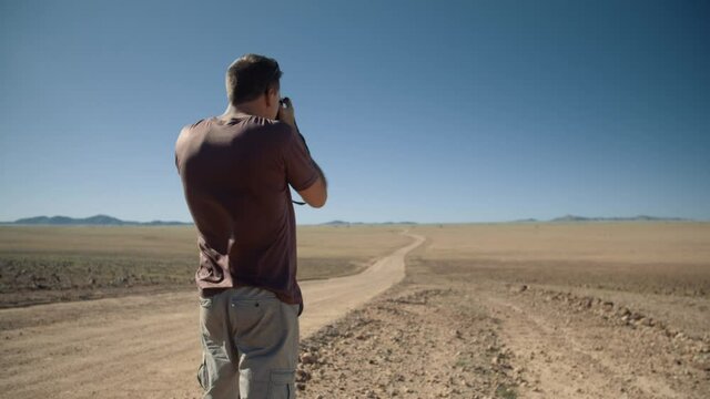 Handheld shot of a photographer standing in a desert taking a landscape photo in Africa as the wind blows on his shirt and a gravel road leading into the distance.