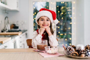 Happy smiling cute little girl kid in Santa Claus hat sitting on the decorated for winter holidays modern kitchen with cocoa drink. Festive season. Waiting for Santa and Christmas gifts. Copy space