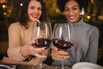 Couple of multicultural girlfriends clebrating toasting red wine at bar restaurant looking at the camera - concept of people drinking alcohol at party